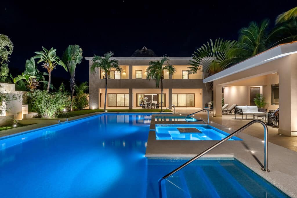 Night view of Marina Palms in Puerto Aventuras featuring an illuminated pool surrounded by lush palms and modern, light-filled two-story building with balconies.