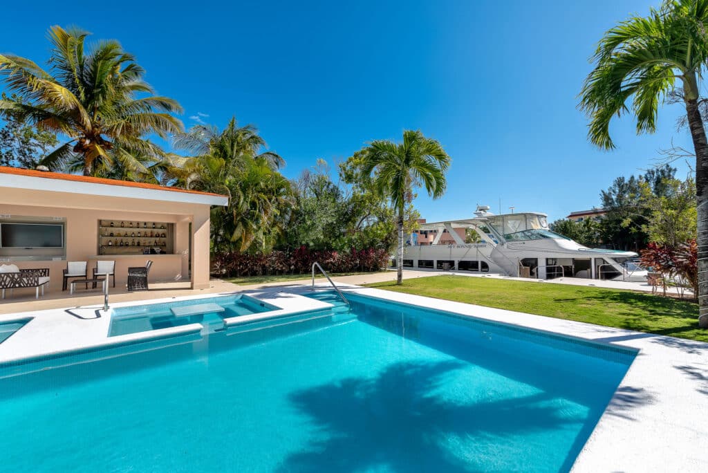 View of a modern outdoor pool area with a boat docked nearby, surrounded by palm trees and lush vegetation at Marina Palms.