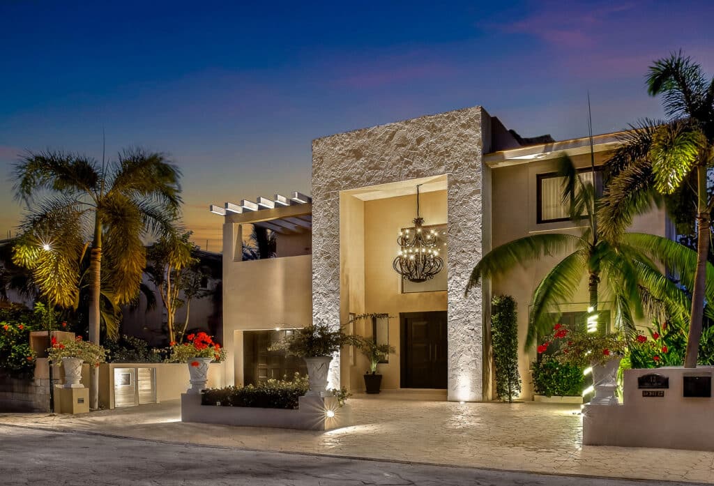 Night view of a luxurious Marina Palms residence in Puerto Aventuras with textured stone walls, a grand chandelier, lush palm trees, and vibrant plants.