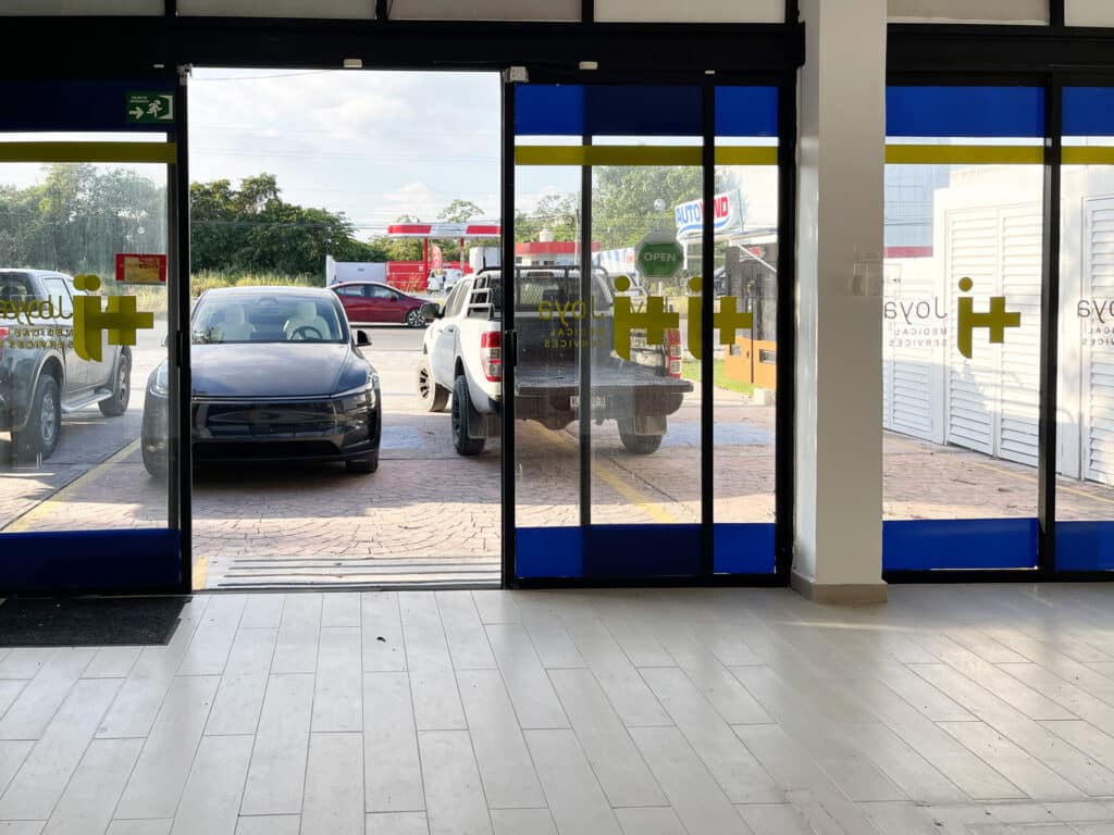 View of vehicles parked outside Prime Highway Commercial Space in Puerto Aventuras through glass doors and windows; the interior has light-colored tile flooring.