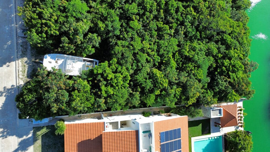A land lot with dense trees located in Phase 4 Canal Front development along the marina in Puerto Aventuras, with a boat visible nearby and residential buildings with red tiled roofs.