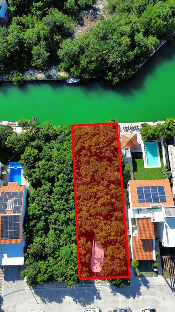 A wooded lot in Phase 4 Canal Front with marina views in Puerto Aventuras, surrounded by residential homes with pools and solar panels, near a canal lined with trees.