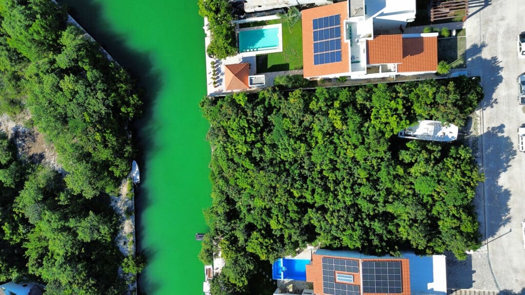 An aerial view of a Phase 4 Canal Front lot in Puerto Aventuras, showing lush green trees along the canal, nearby houses with red-tiled roofs, and solar panels on the rooftops.