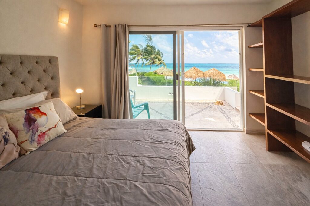 View of a bedroom with a sliding glass door revealing a balcony overlooking a beach with thatched umbrellas, palm trees, and ocean in Puerto Morelos.