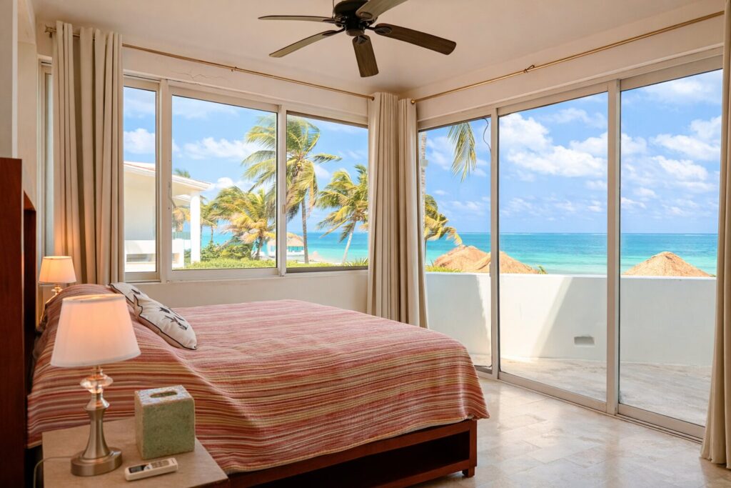 Interior view of a bedroom with large windows overlooking a beachfront scene of palm trees, rocks, the ocean, and blue sky in Puerto Morelos.