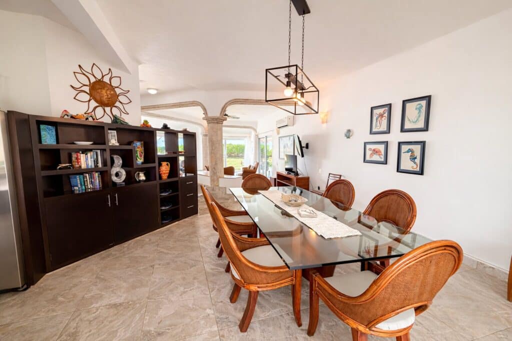 Interior view of Villa Solé Beachfront Home in Puerto Morelos featuring a dining area with a glass table, wooden chairs, and a dark bookshelf against a white wall.