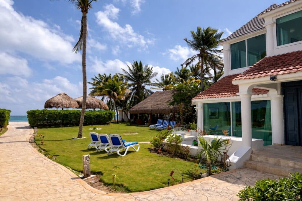 Villa Solé Beachfront Home in Puerto Morelos features a lawn, palm trees, thatched-roof structures, a stone pathway, and ocean views.