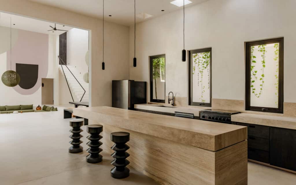 A modern, minimalist kitchen with beige stone counters, black fixtures, and three pendant lights in Villa Coco Dar Lalla in Aldea Zama, Tulum.