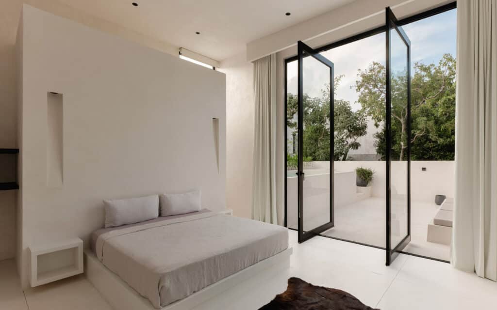 A minimalist bedroom in Villa Coco Dar Lalla in Tulum features a white bed, nightstand, and large glass doors opening to a terrace with greenery and outdoor seating.
