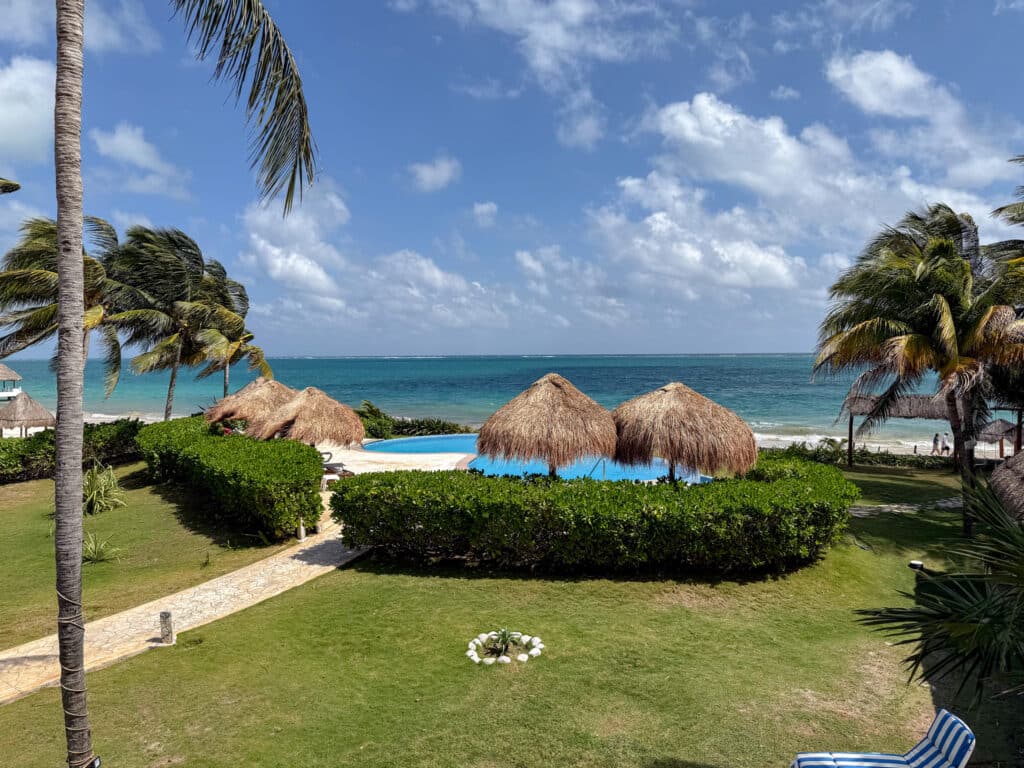 A beachfront view at Villa Solé in Puerto Morelos features a swimming pool with thatched shade structures, surrounded by lush greenery and tall palm trees, overlooking the ocean.