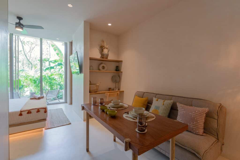 Living area with dining table, sofa, pillows, adjacent to glass doors opening to lush greenery in Aflora L108 loft at Aflora L108 1-Bedroom Loft in Aldea Zama, Tulum.