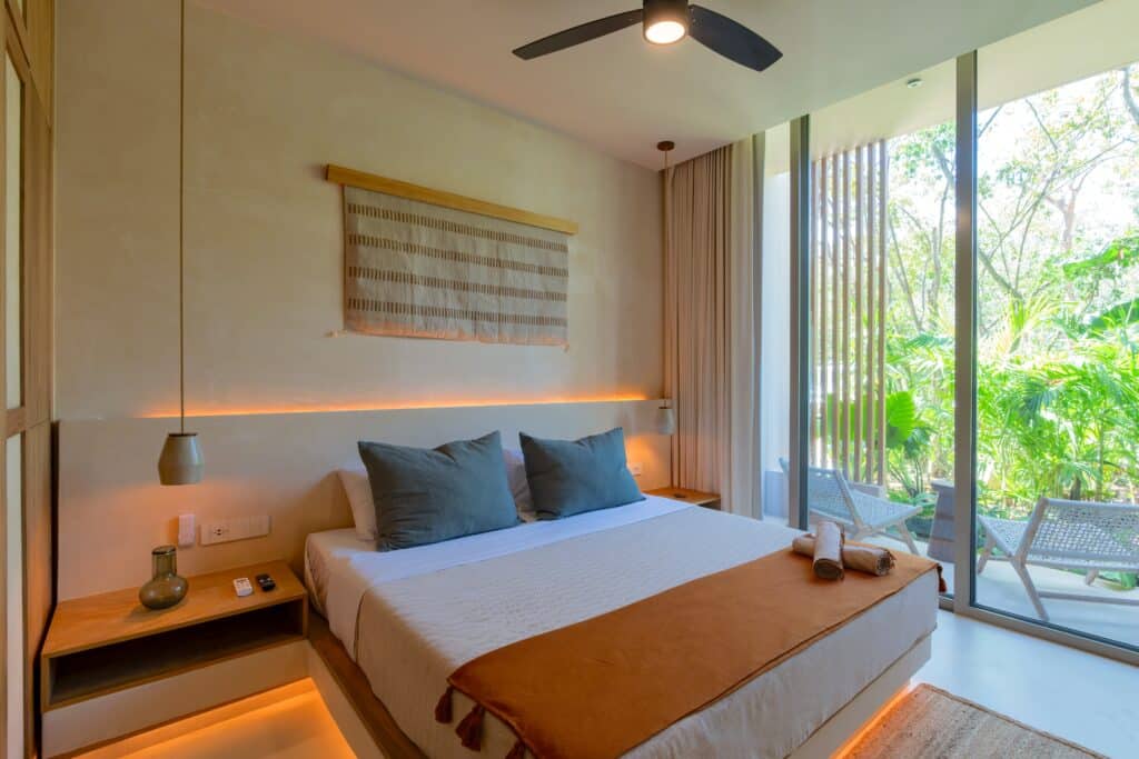 Bright hotel bedroom with a large bed, decorative wall hanging, side table with lamp, and a balcony with greenery and outdoor seating in Aldea Zama, Tulum.