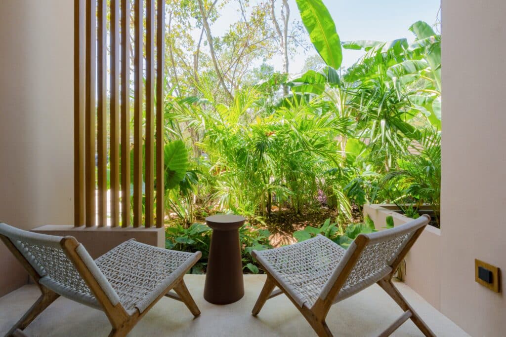 Outdoor patio with wooden slats, surrounded by lush green plants and trees, with seating and a small round table at Aflora L108 1-Bedroom Loft in Aldea Zama, Tulum.