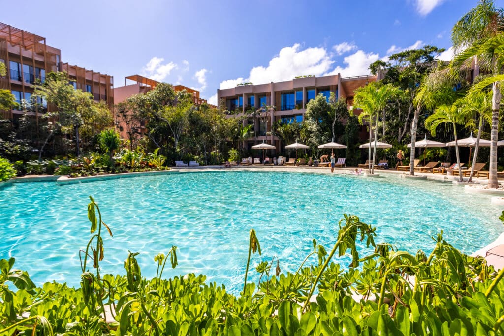 View of a swimming pool surrounded by lush greenery and modern residential buildings at Aflora L108 1-Bedroom Loft in Aldea Zama, Tulum.