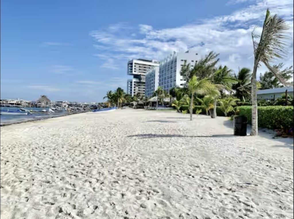 Beachfront view of a sandy shore with palm trees, modern white buildings, and boats docked in the water in Cancun at Amara Beachfront Condo.