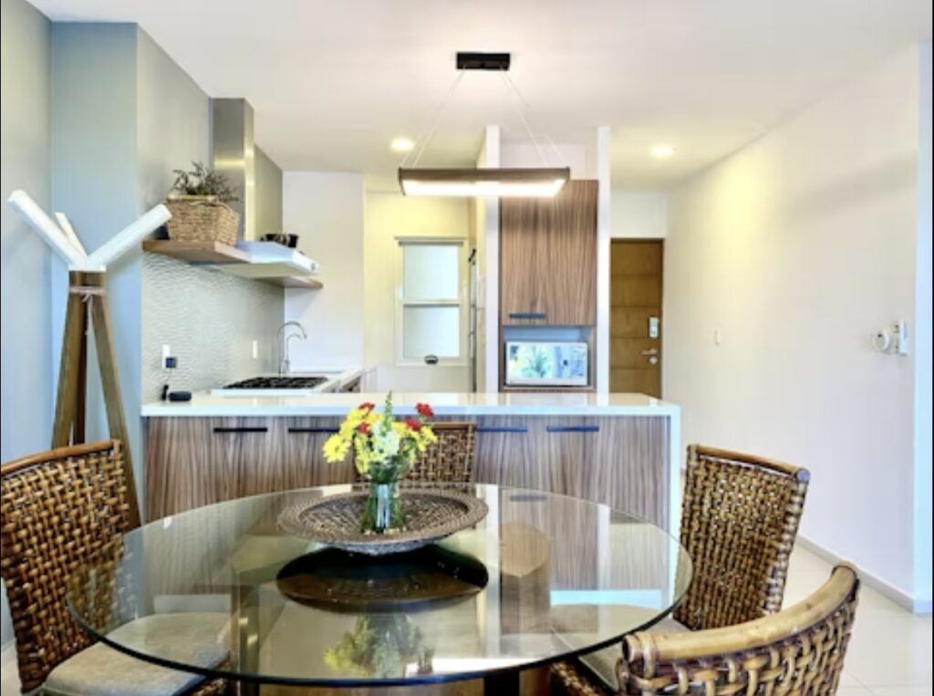 Interior view of a modern kitchen and dining area with a round glass table, wicker chairs, and minimalist decor at Amara Beachfront Condo in Cancun.