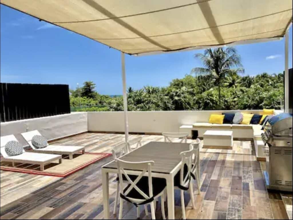 View of a spacious outdoor terrace with lounge chairs, dining table, and lush greenery in the background at Amara Beachfront Condo in Cancun.