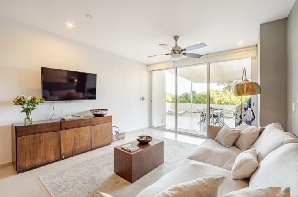 Living room with a white sofa, wooden sideboard, flat-screen TV, and sliding glass doors leading to a balcony overlooking greenery in Beachfront, Cancun.