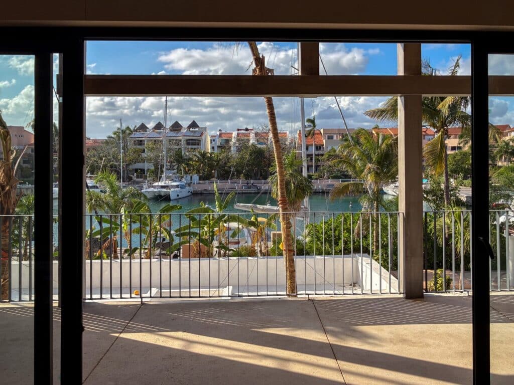 View of a marina with sailboats, palm trees, and residential buildings through a large sliding glass door at Artesan Condo in Caleta Xel - Ha, Puerto Aventuras.