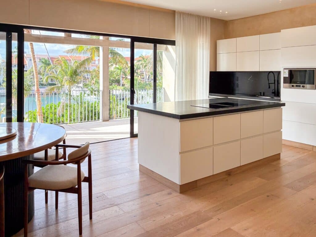 Modern kitchen with white cabinets, black countertops, and large sliding glass doors opening to a tropical view of palm trees and a marina in Caleta Xel - Ha, Puerto Aventuras.