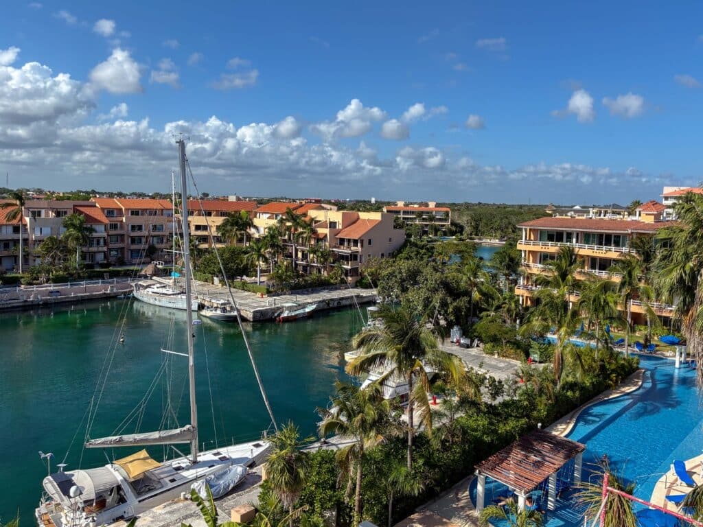 View of a marina with boats, surrounded by multi-story residential buildings, palm trees, and a swimming pool on a sunny day at Puerto Aventuras at Artesan Condo.