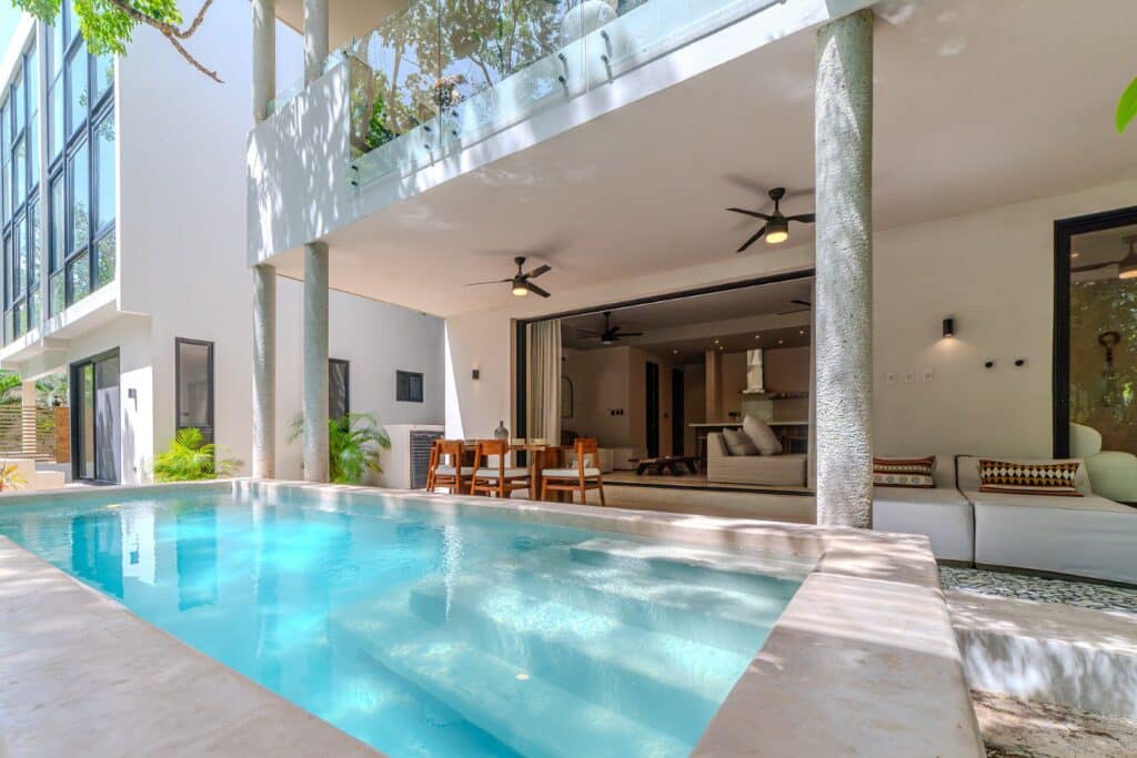 Open living area with large glass sliding doors, outdoor pool, modern furniture, ceiling fans, and lush greenery at property in Holistika, Tulum.