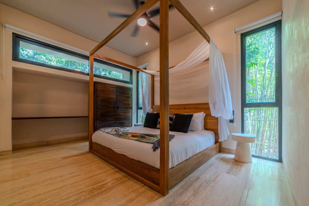 Bedroom with a wooden four-poster bed, large windows with greenery view, and light-colored flooring in a natural-style interior at Holistika, Tulum at Atta Holistika A1.