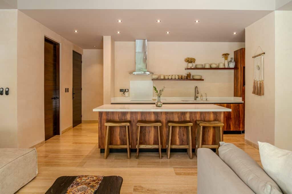 Modern kitchen with wooden cabinetry, a white countertop island, open shelving, and a stainless steel range hood in a contemporary living space in Holistika, Tulum.