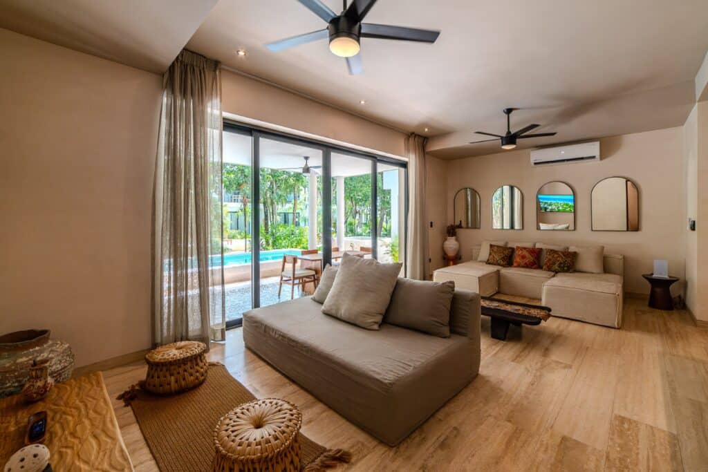 Living room with beige sofas, wall mirrors, and view of patio and pool through large glass doors in Holistika, Tulum.