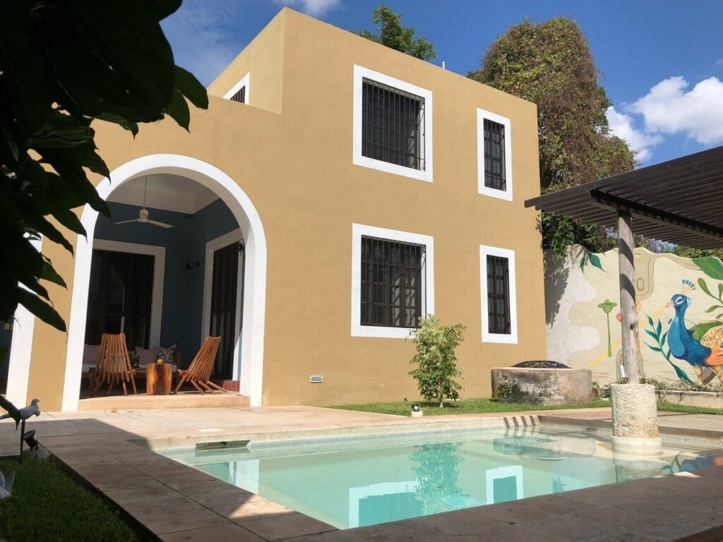 Bright yellow two-story house with white-framed windows, a covered patio with outdoor chairs, a small swimming pool, and garden in Centro Histórico, Merida.