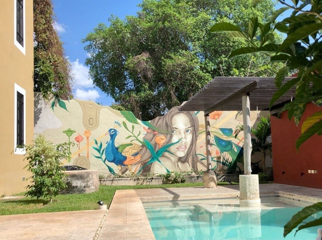 Colorful mural of a woman's face with birds and nature elements on a wall behind a pool in a courtyard near trees at Barrio De Santiago Villa in Centro Histórico, Merida.