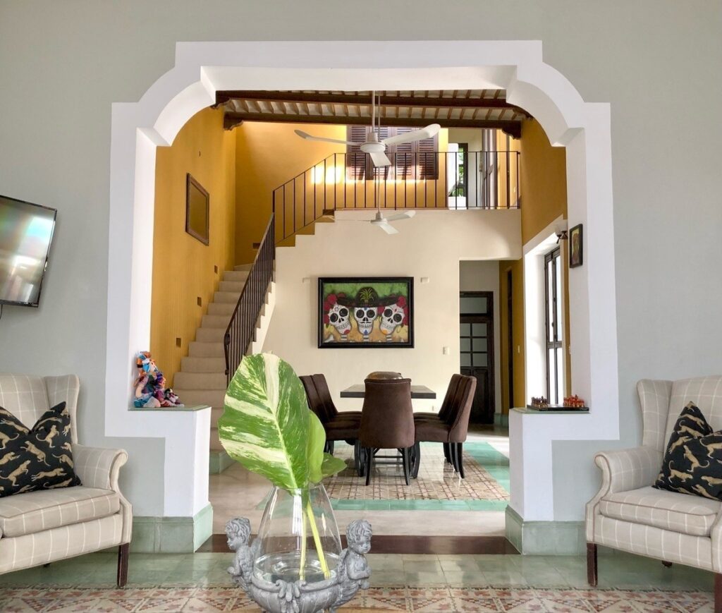 Interior view of a living room with beige sofas, a dark dining table with four chairs, artwork, and an arch doorway leading to a staircase in Centro Histórico, Merida.