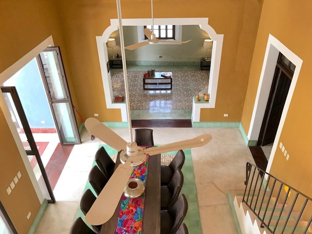 Interior view of a two-story home with yellow walls, a wooden dining table, black chairs, and a ceiling fan, located at Barrio De Santiago Villa in Centro Histórico, Merida.