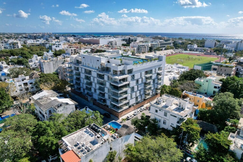 Modern white and gray condominium building with balconies, surrounded by trees and neighboring structures at Ceiba at 25 2-Bedroom Condo in Playa Centro, Playa del Carmen.