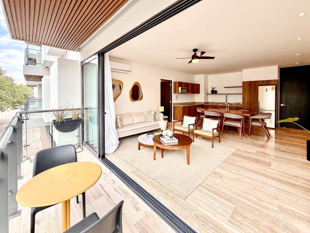 Modern living room with sliding glass doors opening to a balcony, electric fan, wooden furniture, and open kitchen in Playa Centro at Ceiba at 25 2-Bedroom Condo.