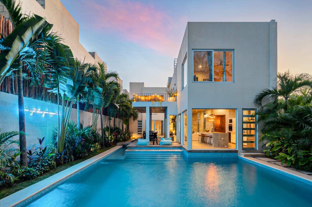 Modern white two-story villa in Tulum with large glass windows, outdoor pool, and tropical landscaping, showcasing a contemporary design.