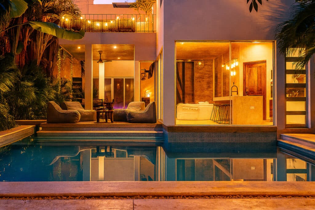 Nighttime view of Villa Cielo Azul in Tulum with an illuminated outdoor living space, large glass windows, and a reflecting pool surrounded by lush tropical plants.