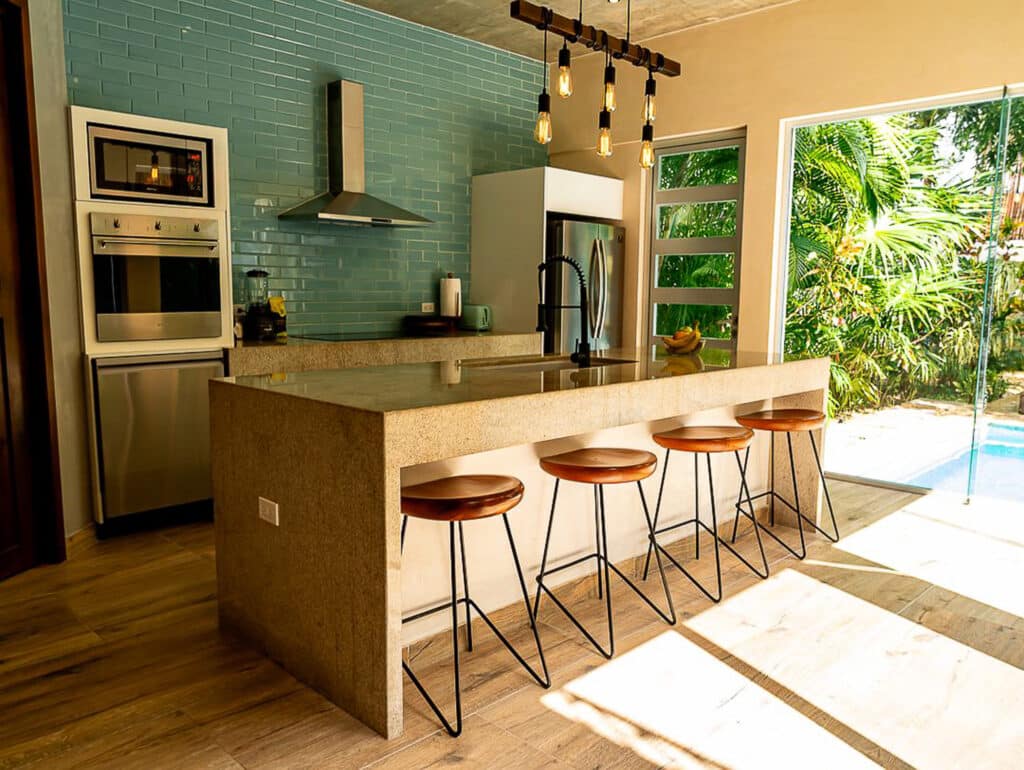 Modern kitchen in Villa Cielo Azul, Tulum, featuring a green tiled backsplash, beige stone island with bar stools, stainless steel appliances, and large glass doors opening to lush greenery.