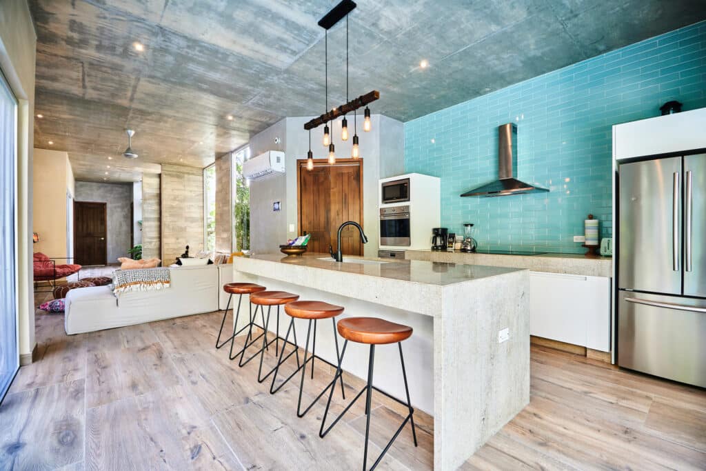 Interior of Villa Cielo Azul in Tulum features a modern kitchen with turquoise tile backsplash, concrete ceiling, and open-plan living area with wooden flooring.