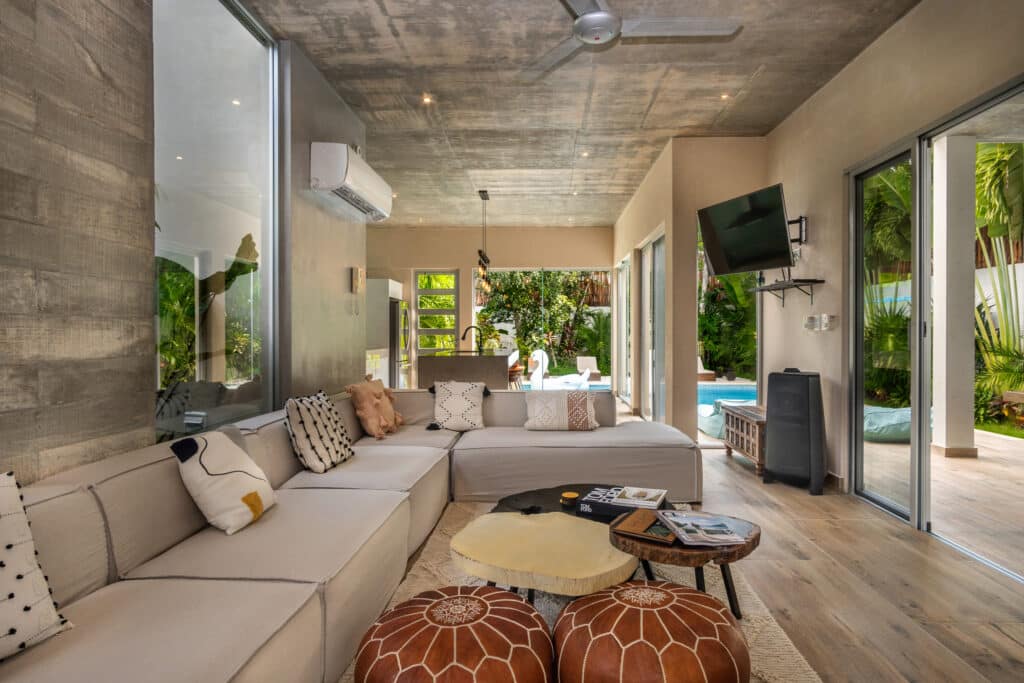 Interior of Villa Cielo Azul in Tulum showing a modern living area with a large beige sectional sofa, wooden flooring, and floor-to-ceiling glass doors opening to a lush outdoor space.
