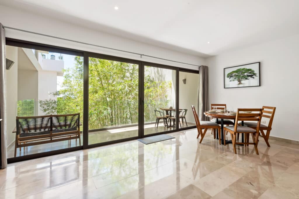 Living room with sliding glass doors opening to a balcony with greenery, dining area with wooden table and chairs, and a framed tree artwork on white wall.