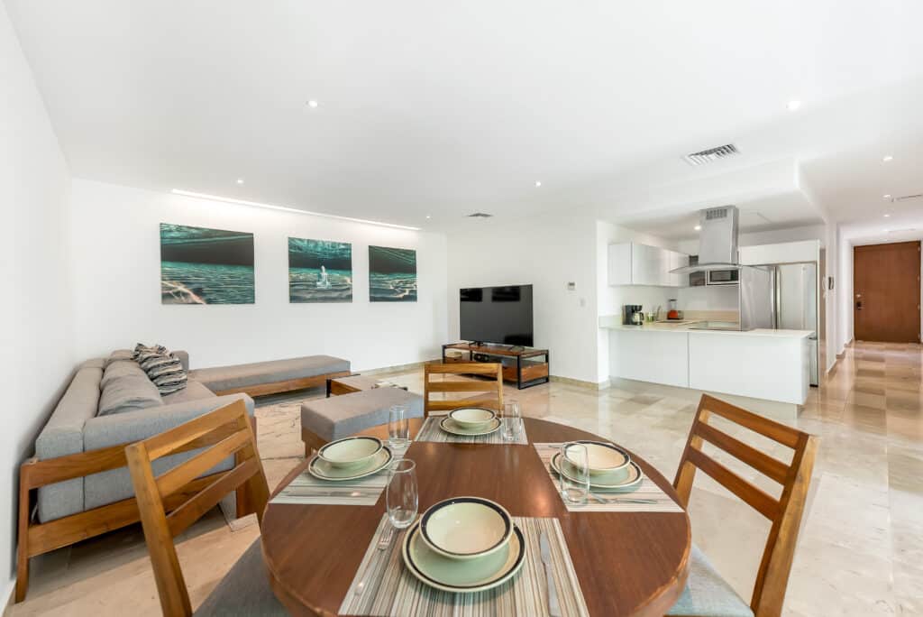 Living room with a gray sofa, wooden dining table with place settings, wall art, flat-screen TV, and open kitchen in a modern apartment in Playa Centro, Playa del Carmen.