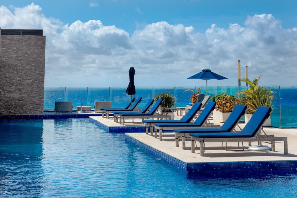 Poolside area with lounge chairs, umbrellas, and potted plants overlooking the ocean against a blue sky with clouds in Playa Centro, Playa del Carmen.