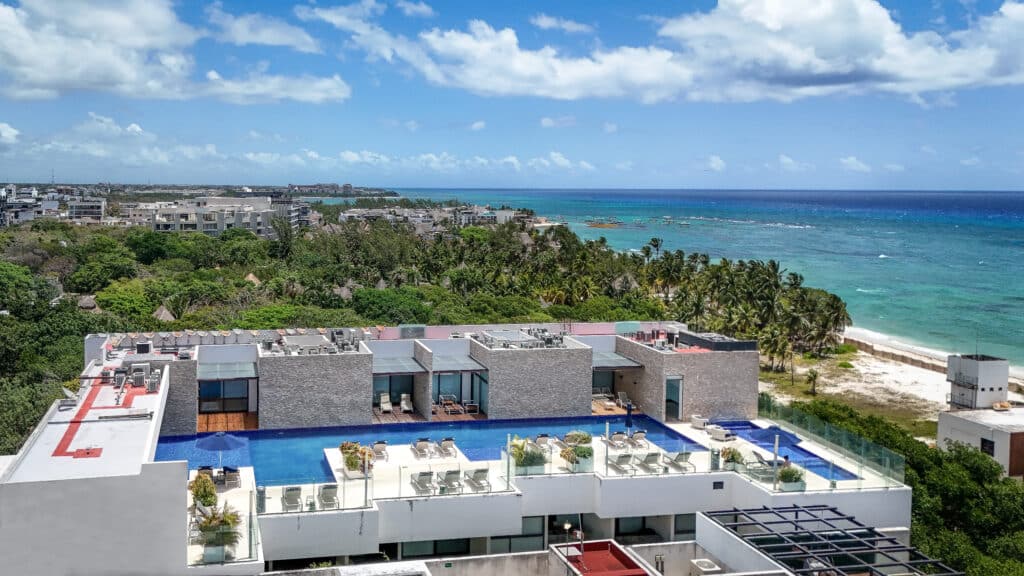 View of the ocean and sandy beach from a high-rise condo rooftop in Playa Centro, Playa del Carmen, with a swimming pool and lush greenery at Cruz con Mar 407 2-Bedroom Condo.