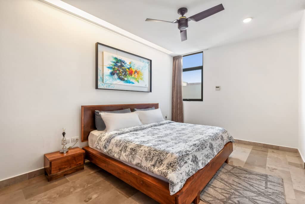 Bedroom with a wooden bed, patterned bedding, two bedside tables, modern lamp, abstract artwork, window with curtain in Playa Centro, Playa del Carmen.