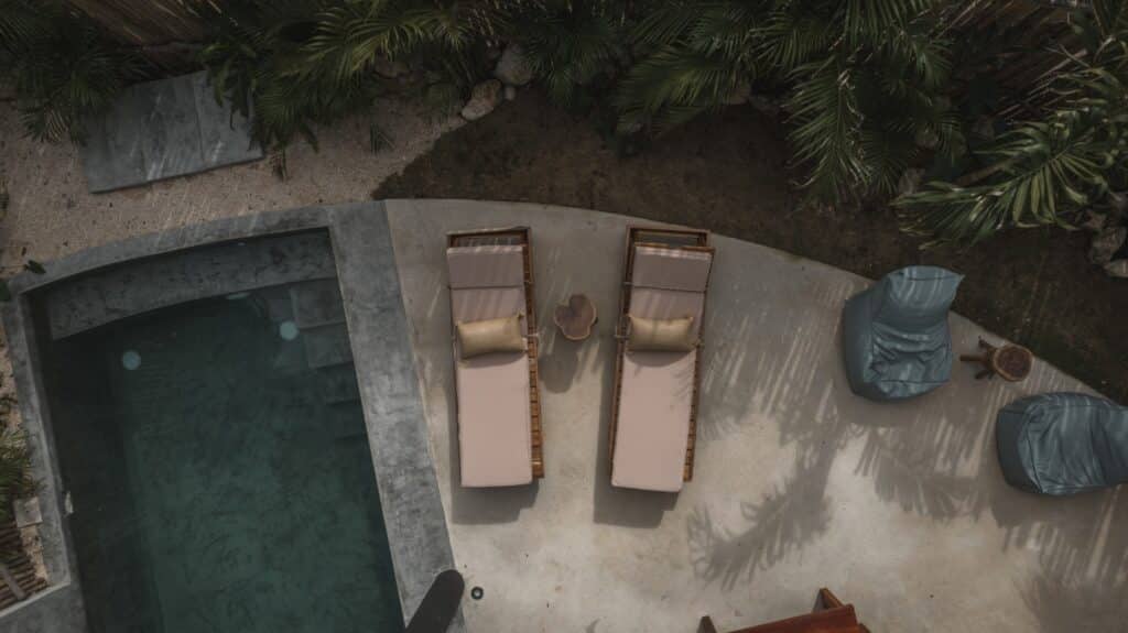 Top-down view of two beige lounge chairs and a small table on a curved concrete patio next to a swimming pool, surrounded by tropical plants at Esencia Villa in La Veleta, Tulum.