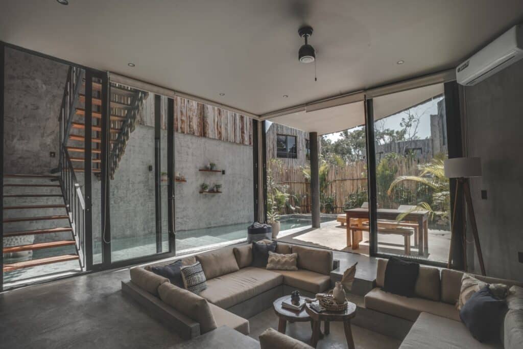 Interior of a modern living room with concrete walls, large glass doors opening to a private pool and outdoor seating area at Esencia Villa in La Veleta, Tulum.