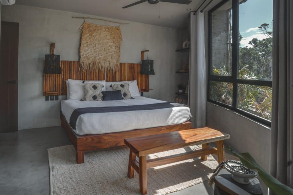 Bedroom with a large bed, wooden headboard, and decorative pillows, near a window with views of trees at Esencia Villa in La Veleta, Tulum.