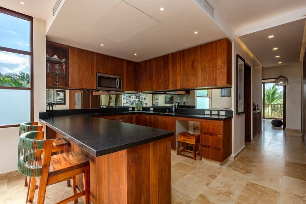 Interior view of a kitchen with wooden cabinets, black countertops, and a large window overlooking tropical trees in a beachfront property at Kaoba Beachfront Condo.