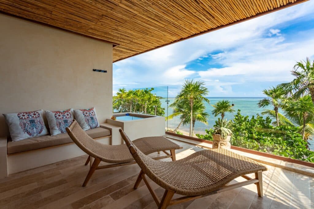 Balcony with wicker lounge chairs, lush green palm trees, and ocean view overlooking the sea at beachfront, Tulum at Kaoba Beachfront Condo.